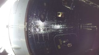 An astronaut can be seen smiling in the window of a dark spacecraft capsule as light reflects off its outer hull.