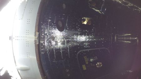 An astronaut can be seen smiling in the window of a dark spacecraft capsule as light reflects off its outer hull.