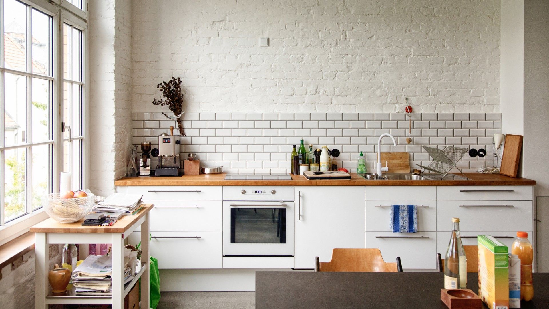 a white oven in a large, airy white kitchen with a brick wall, a wall-mounted countertop, and a dining table in the middle full of kitchen items