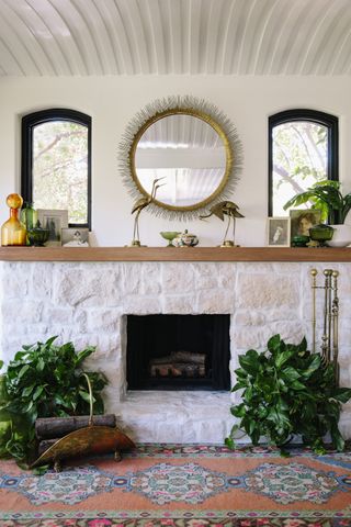 A white stone fireplace with two windows and a round mirror above the mantel
