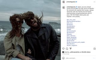 Jason Momoa and Adria Arjona smiling together with their hair blowing in the wind while standing on a beach.