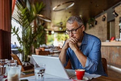 A retired man looks at his laptop in a cafe, appearing worried.