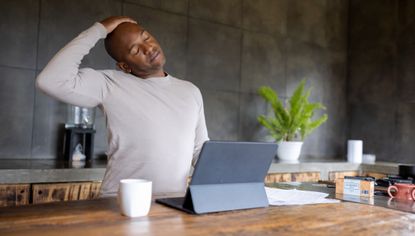 Man sitting at kitchen island stretches his neck to the side. A tablet is propped up in front of him.