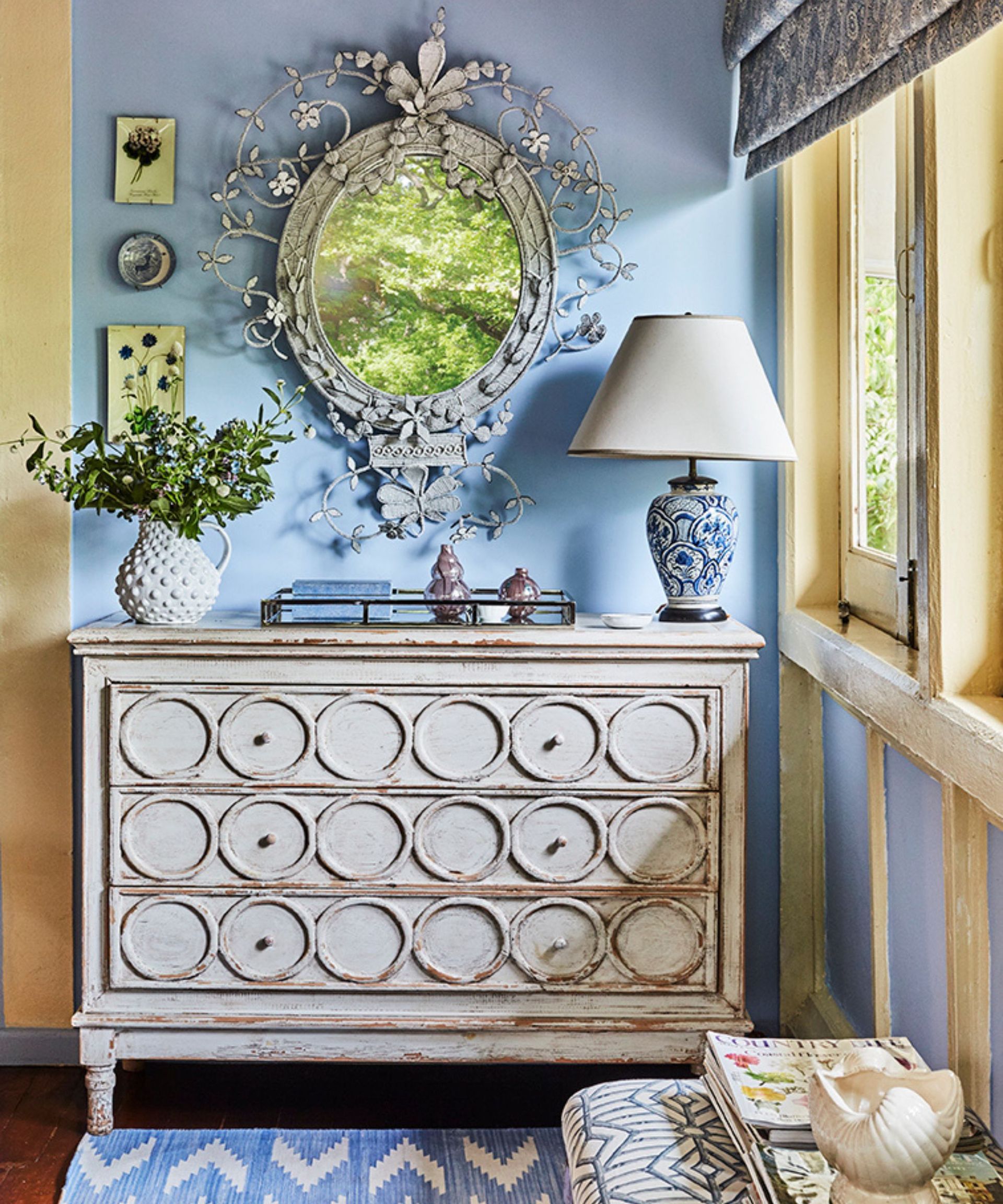Cornflower blue wall with ornate floral gray mirror on wall. There is a white washed dresser with drawers, chevron blue rug and bright windows with yellow frames.
