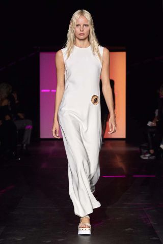 A female model wearing a long sleeveless white dress walking down a runway.