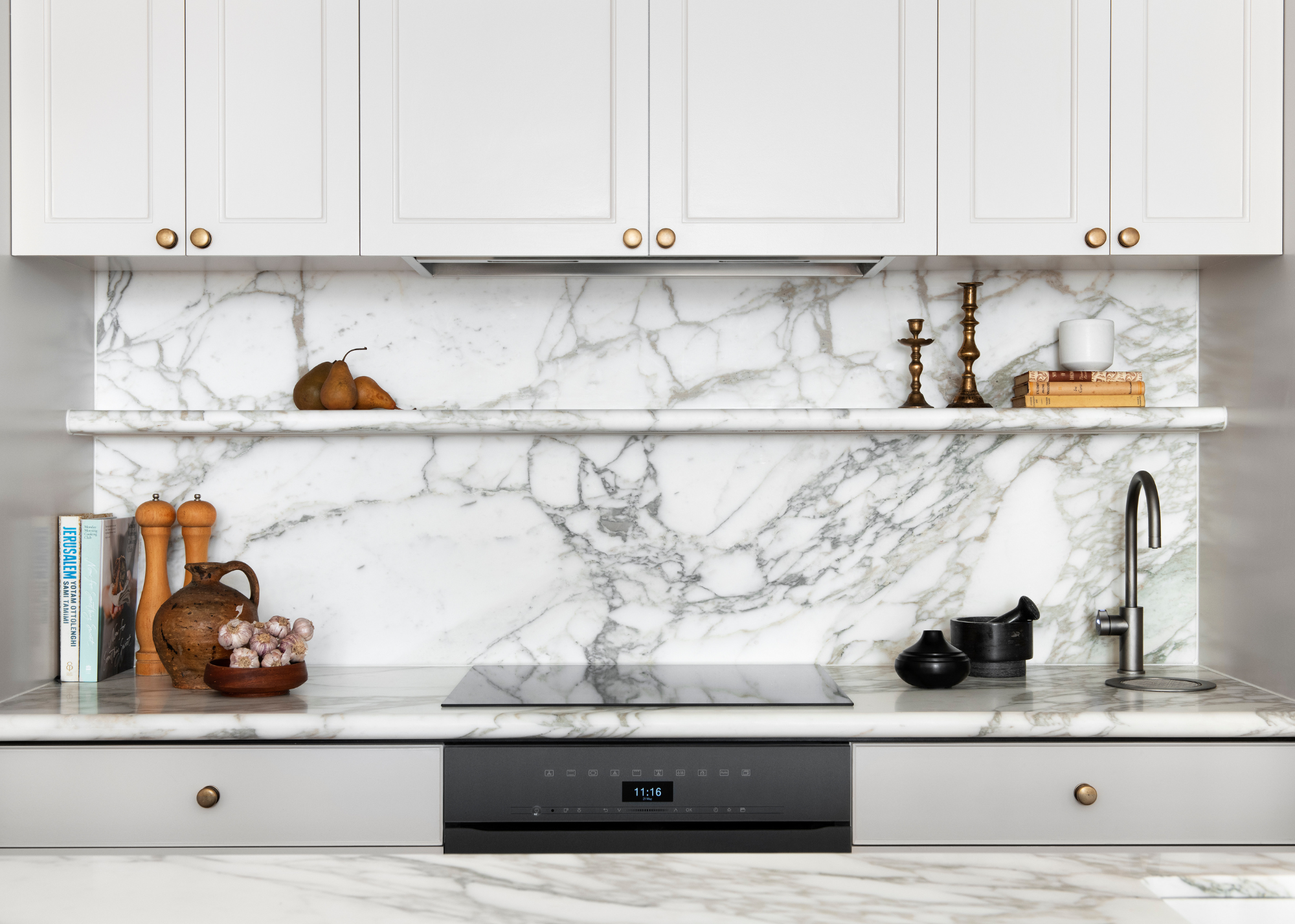 A white kitchen with a stone backsplash, brass candlesticks, pepper and salt mills, a bowl of garlic, and a mortar and pestle