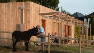 Man standing in front of his self build home next to a field with a donkey. His wife sits in the background on a sunny veranda