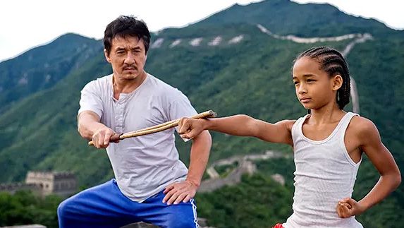 A still from the 2010 movie The Karate Kid in which Jaden Smith is playing Dre Parker and punching and Jackie Chan is playing Mr. Han stood next to him and training him.