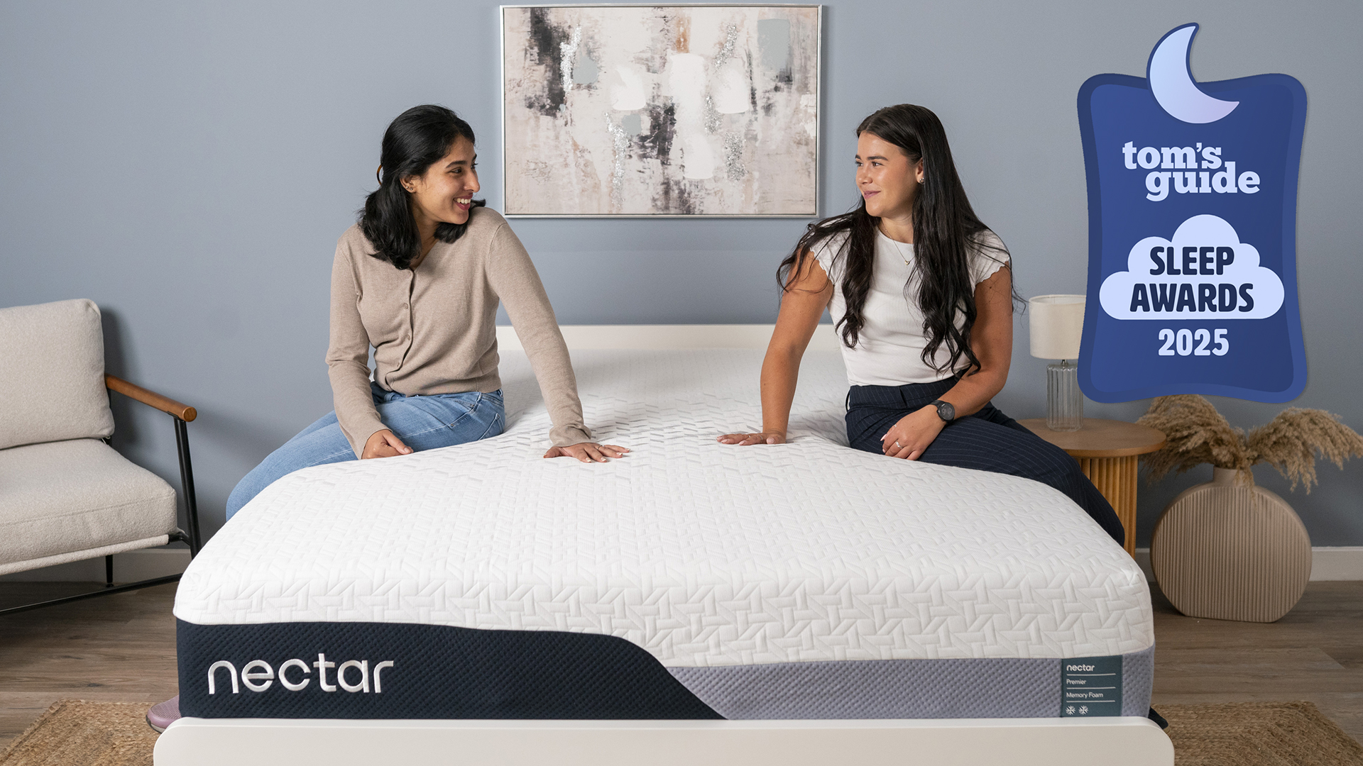 This image shoes two of our testers sitting on the Nectar Premier Memory Foam mattress on a bed frame in a decorated bedroom. On the left is Becky George, on the right is Eve Davies. They both sit on the edge of the mattress, testing the support and comfort. In the top right corner is a Tom&#039;s Guide Sleep Awards 2025 graphic (the Nectar Premier has won best memory foam mattress)