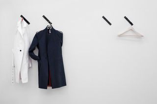 View of ’Murder Weapons’ by Matthew Brannon and Carlo Brandelli featuring a white coat and a blue coat on black individual, wall-mounted coat hooks on a light coloured wall