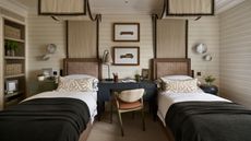 a twin childrens bedroom with beige striped walls and two canopy twin beds, a desk in the middle, a bookcase, and dark green blue color pops