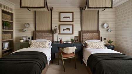 a twin childrens bedroom with beige striped walls and two canopy twin beds, a desk in the middle, a bookcase, and dark green blue color pops