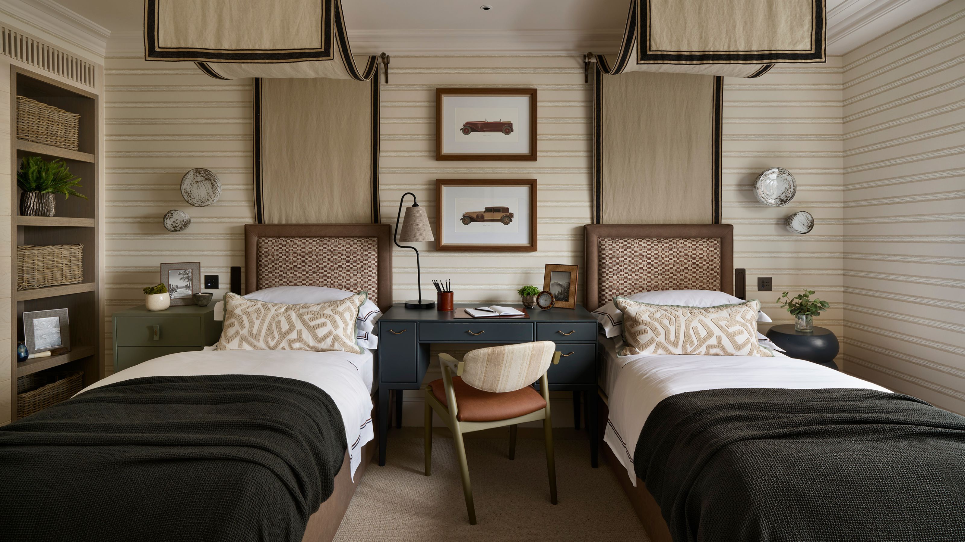 a twin childrens bedroom with beige striped walls and two canopy twin beds, a desk in the middle, a bookcase, and dark green blue color pops