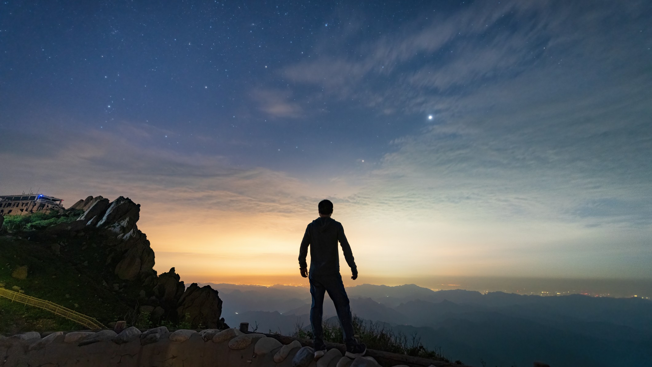 a man silhouetted looking out across mountainous landscape and the predawn sky.