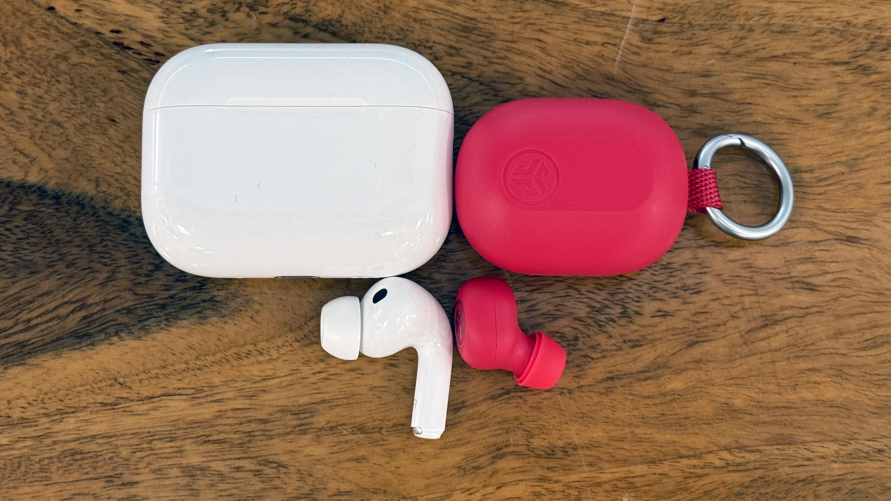 JLab JBuds Mini ANC and AirPods Pro 3 next to each other, showing how much smaller the earbud and case are compared to AirPods