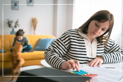 mother managing household budget on calculator at table while child plays on sofa with a dog