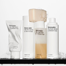 Five Nécessaire body products with soap bubbles and water droplets running down them stand on a black shelf against a white wall.