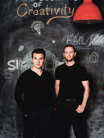 Two males with short black hair and black clothing standing against a chalkboard. 