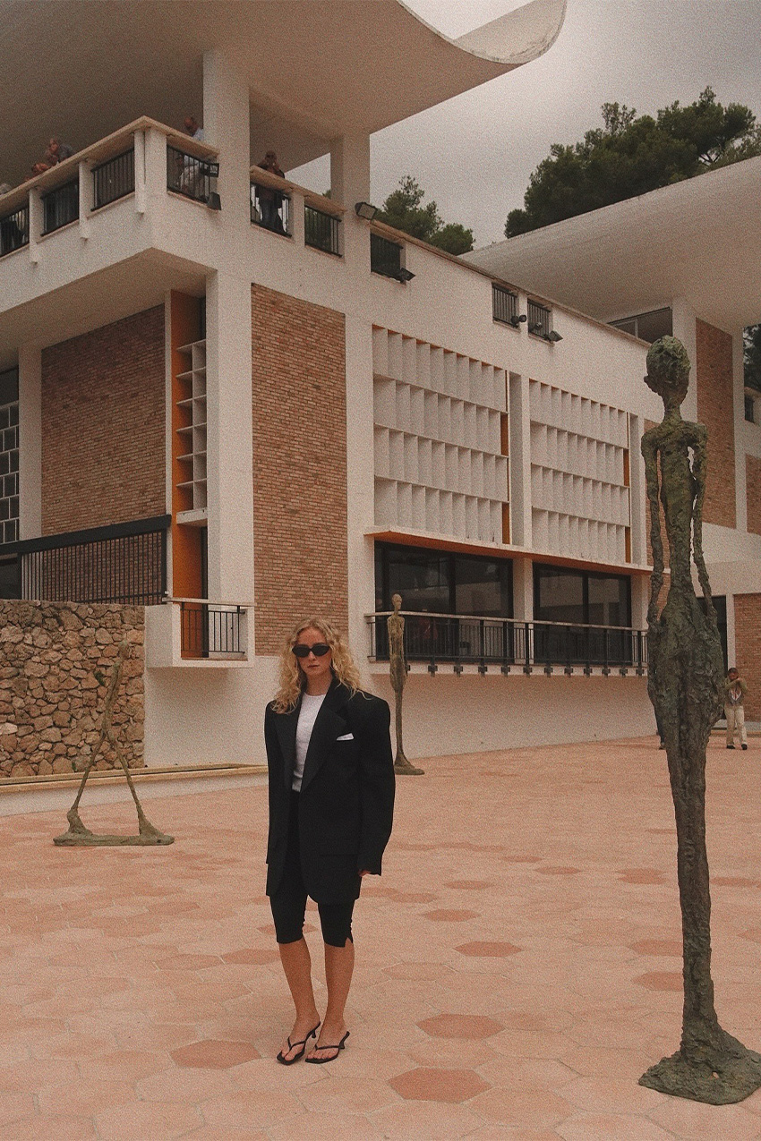 European vacation outfit idea is shown in a photo of a woman at a museum wearing black sunglasses with a black boxy blazer, white t-shirt, black capri pants, and heeled thong sandals