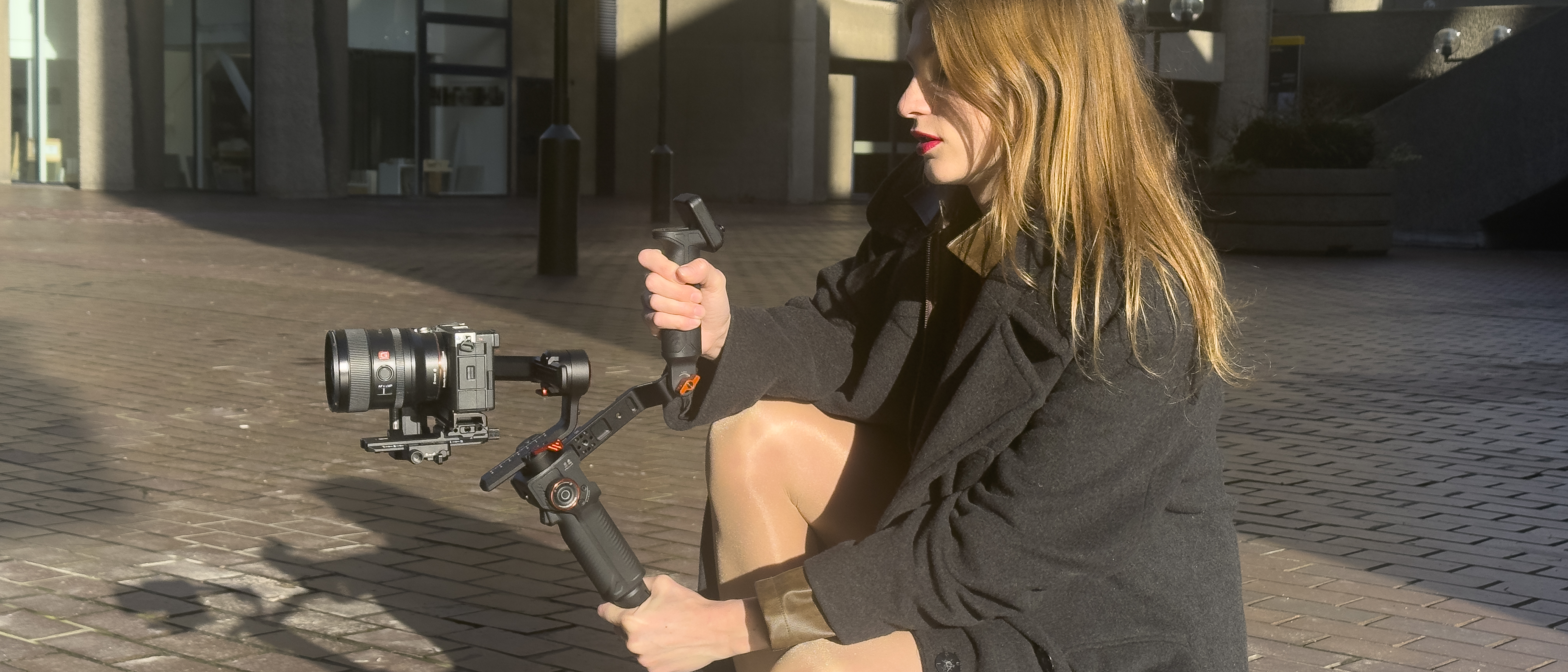 Person holding a Hohem MT3 gimbal stabilizer with a Sony mirrorless camera attached while crouching outdoors on a paved plaza.