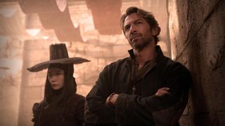 Still from the movie Rebel Moon (2023). Here we see Nemesis (a woman dressed in black wearing a wide-brimmed black hat) and Gunnar (adult male with short swoopy hair and a beard, wearing all black) standing together.
