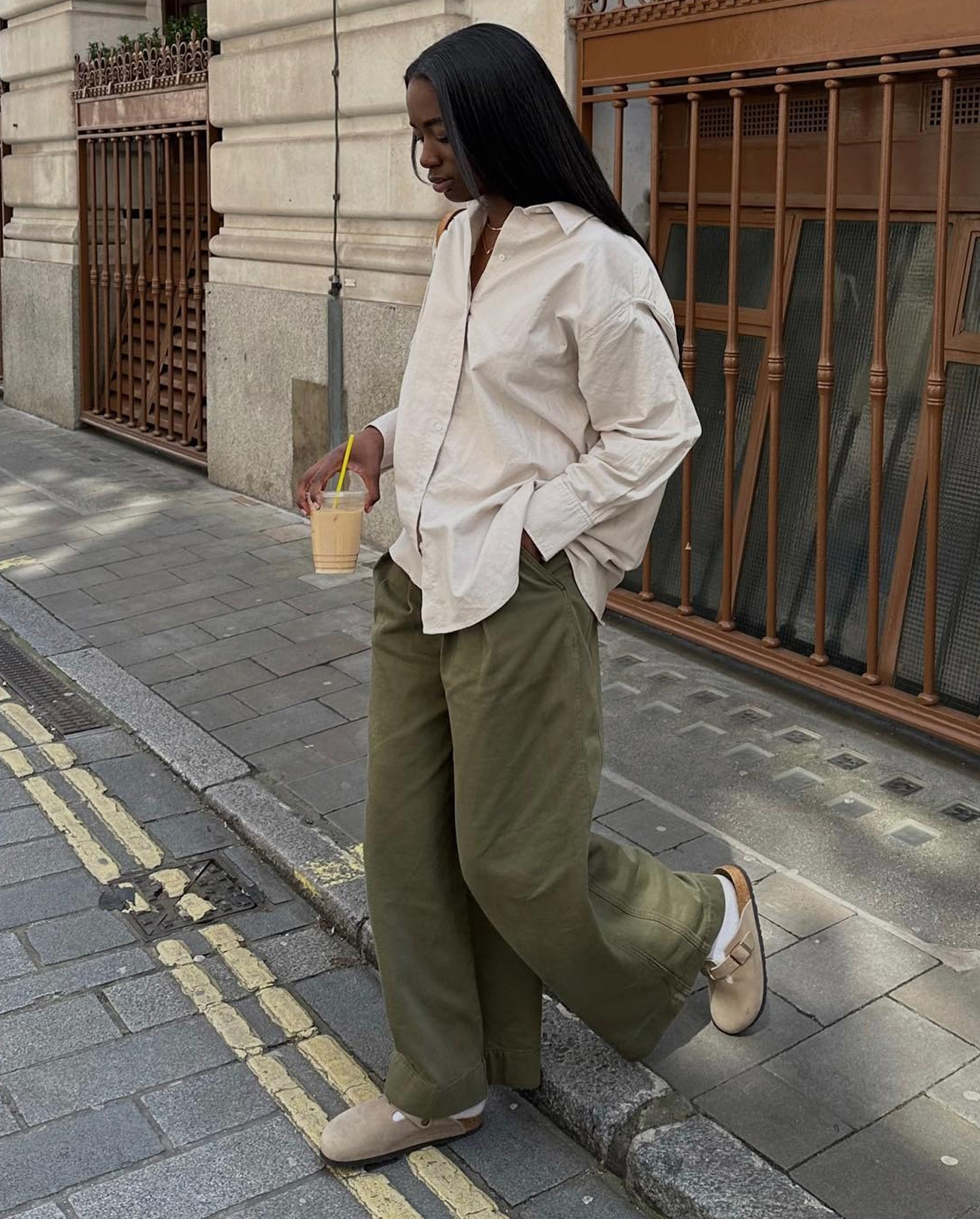 British style influencer Liv Madeline poses on a sidewalk wearing an oversized beige button-down shirt, wide-leg green pants, white socks, and tan Birkenstock Boston clogs