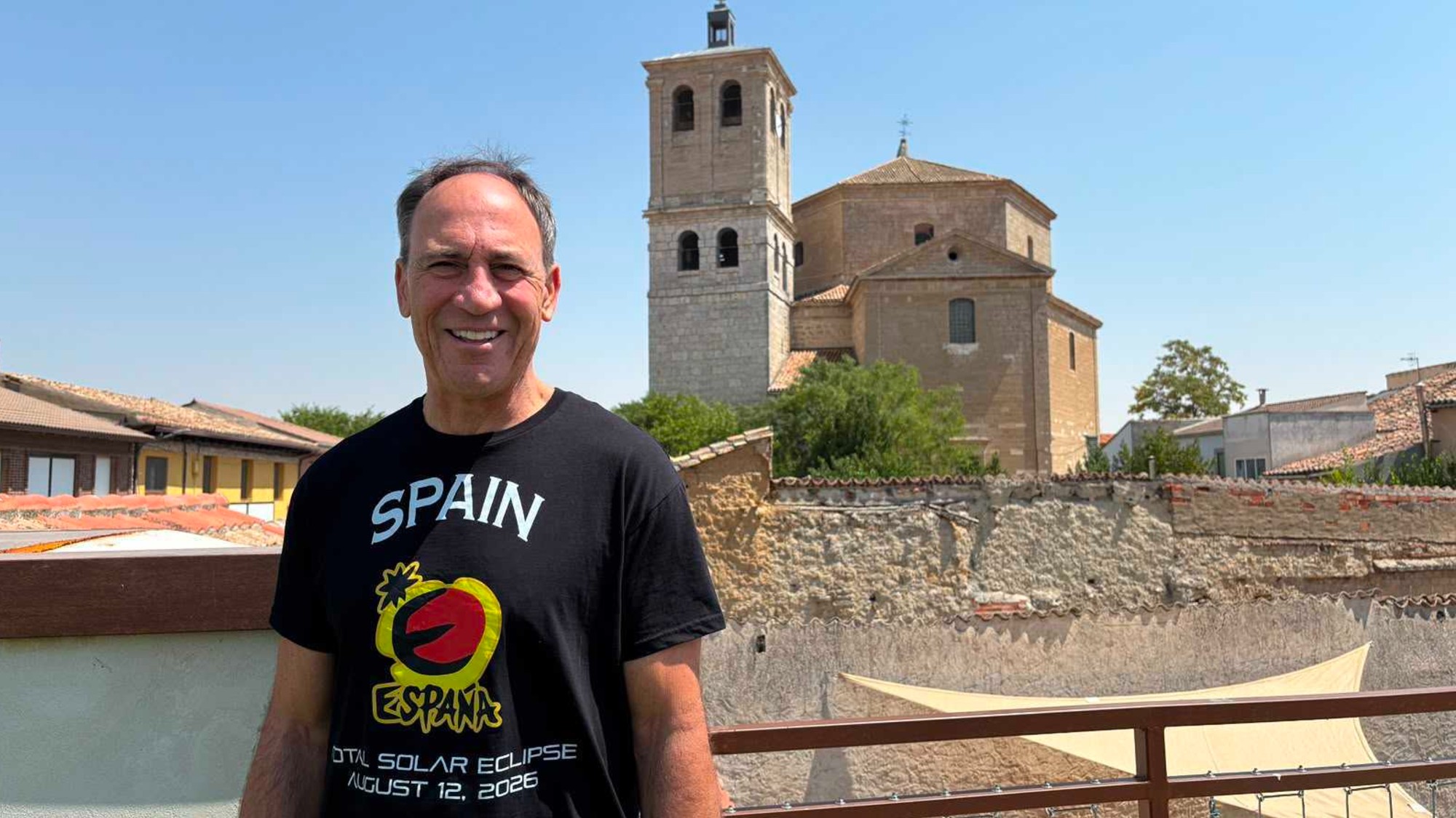 a man in a t-shirt that reads 'spain total eclipse 2026' stands in front of a stone church