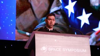 a man in a black military uniform speaks into microphones at a lectern on a stage lit in blues and purples