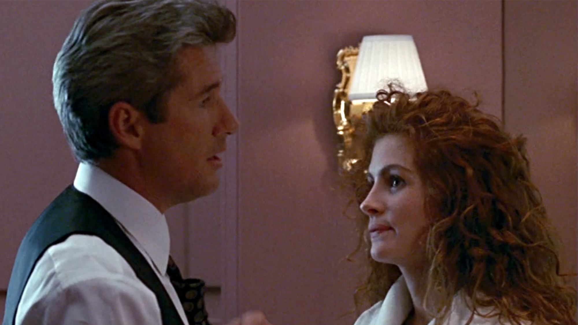 Richard Gere on the left, tying a tie with Julia Roberts in a robe, looking at him.