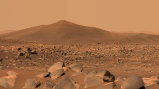 a rust orange yellow sky and landscape features a rocky surface in the foreground, with a large pointed hill beyond.