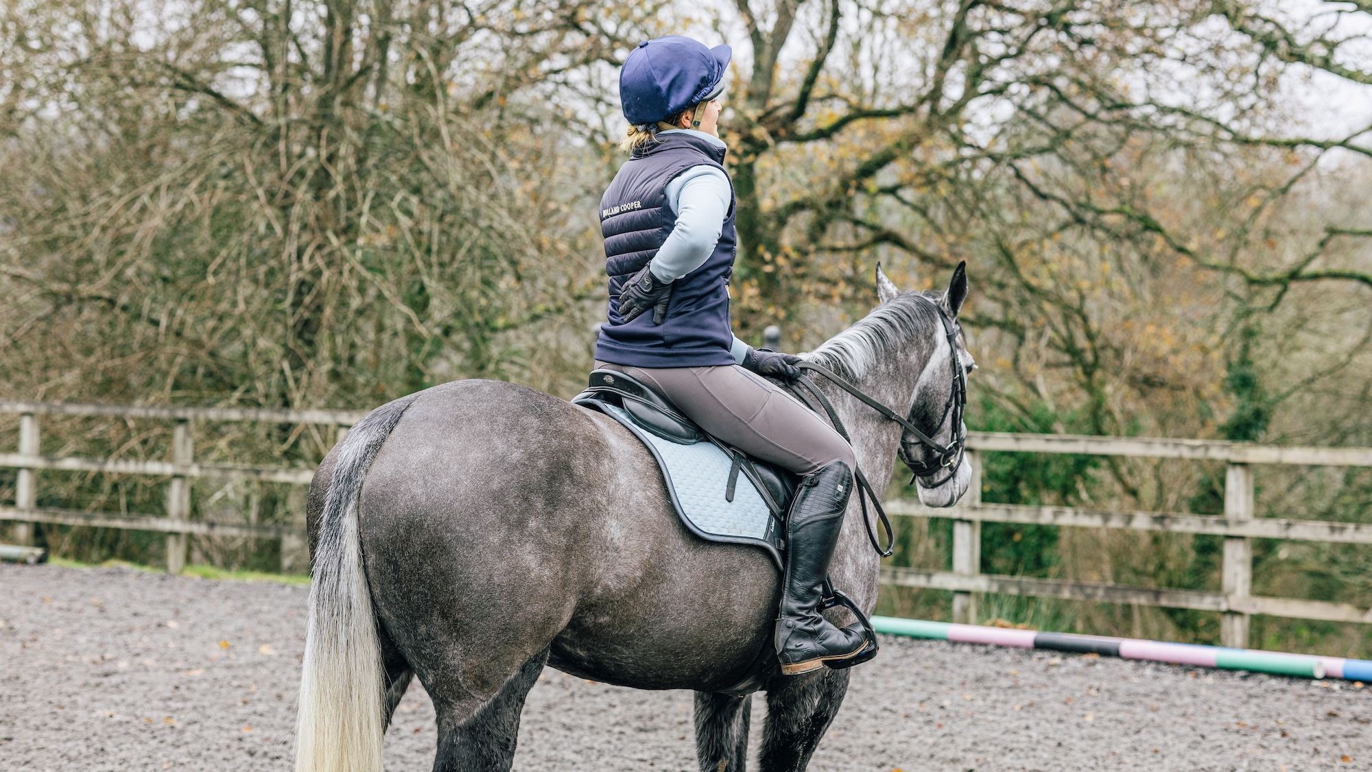 Rider in a blue gilet holds her back with her right hand, while sitting on a dark grey horse in an all-weather arena.