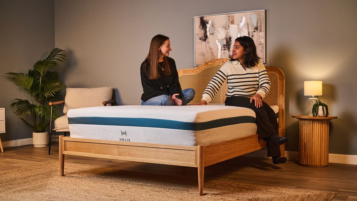 Two women sit on the Helix Twilight mattress on a bed frame in a bedroom, testing the motion isolation, comfort and support