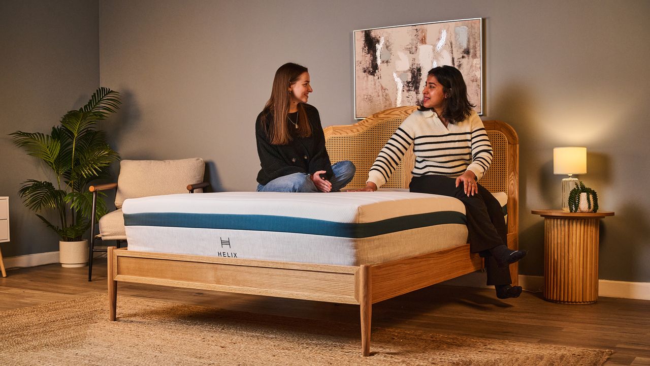 Two women sit on the Helix Twilight mattress on a bed frame in a bedroom, testing the motion isolation, comfort and support
