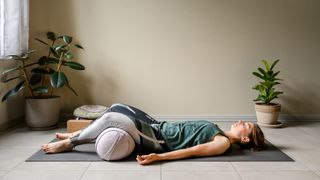 Woman lying on a yoga mat with a cushion under her knees with two potted green plants on either of her side.