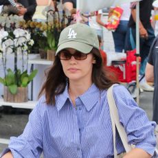 Lucy Hale wearing a white striped button down, gray jeans, nude sandals, and a tote bag, and a green LA cap