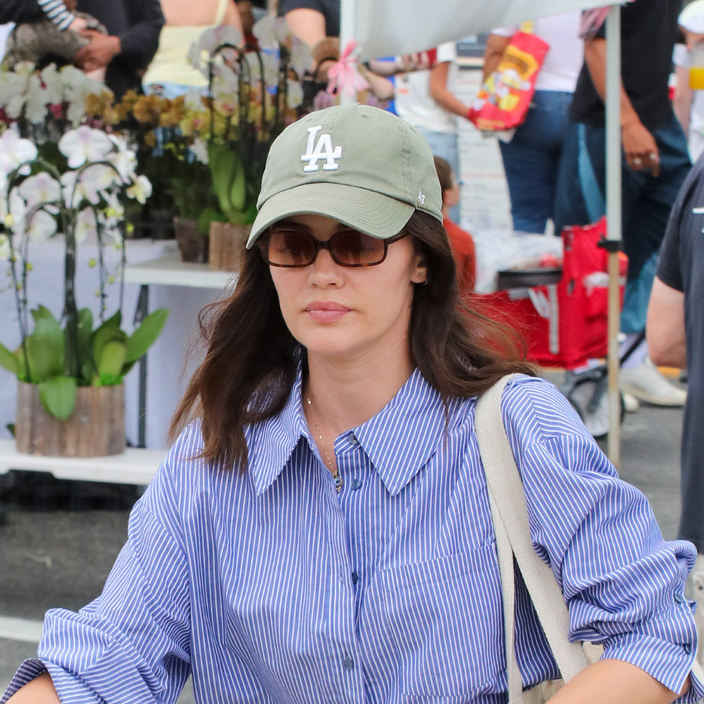 Lucy Hale wearing a white striped button down, gray jeans, nude sandals, and a tote bag, and a green LA cap