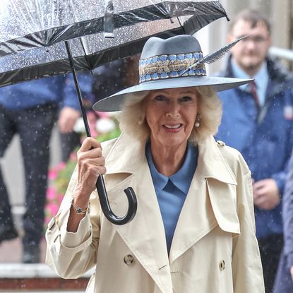 Queen Camilla holding an umbrella wearing a trench coat and hat