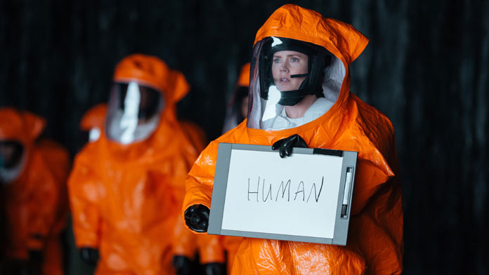 A still from the movie Arrival in which Amy Adams' character Louise is wearing an orange hazmat suit and holding a sign that says HUMAN.