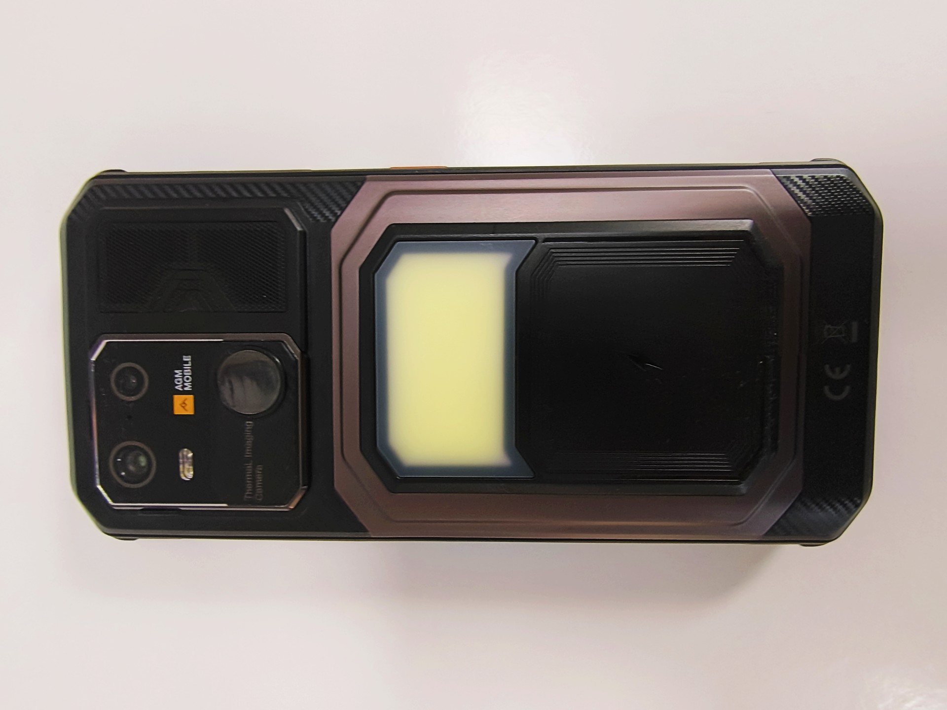 An AGM G3 Pro rugged smartphone with a thick, protective black casing and visible app icons on its screen is shown lying on a white surface next to a much thinner green smartphone.