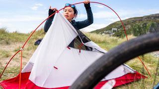 A woman is putting up a MSR Hubba Hubba Bikepack One-Person Tent on a patchy of sandy grass. There is a bicycle wheel in the foreground.