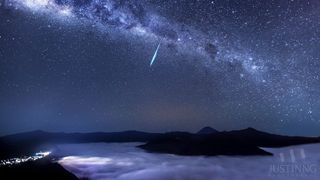 The Milky Way stretches across the sky and a bright Eta Aquarid meteor streaks across the center. Below is the top of Mount Bromo surrounded by clouds. 