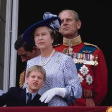 Young Prince William has dark blonde hair and wears a suit, while posing with Queen Elizabeth II on Buckingham Palace balcony