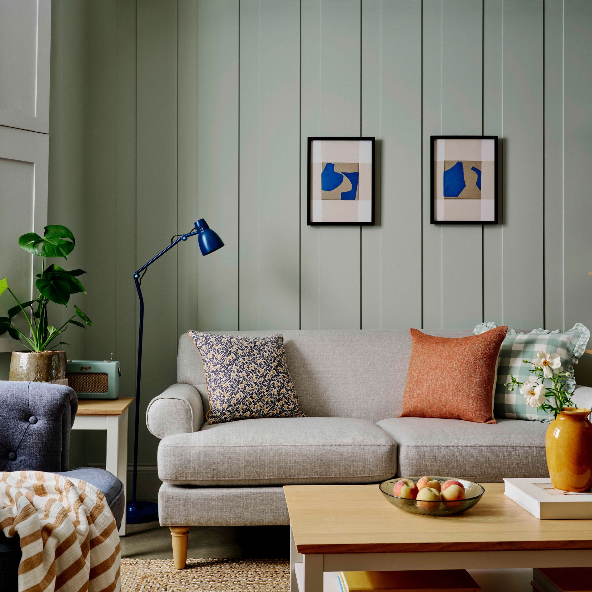 cottagecore living room ideas, living room with panelled walls painted in a pale green, neutral sofa, wooden coffee table, blue floor lamp, artwork, navy armchair, jute rug, orange cushions, plant