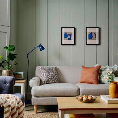 cottagecore decor ideas for living rooms, living room with panelled walls painted in a pale green, neutral sofa, wooden coffee table, blue floor lamp, artwork, navy armchair, jute rug, orange cushions, plant 