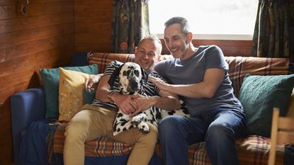 A same-sex couple cuddle with their dog on the sofa at a vacation cabin.