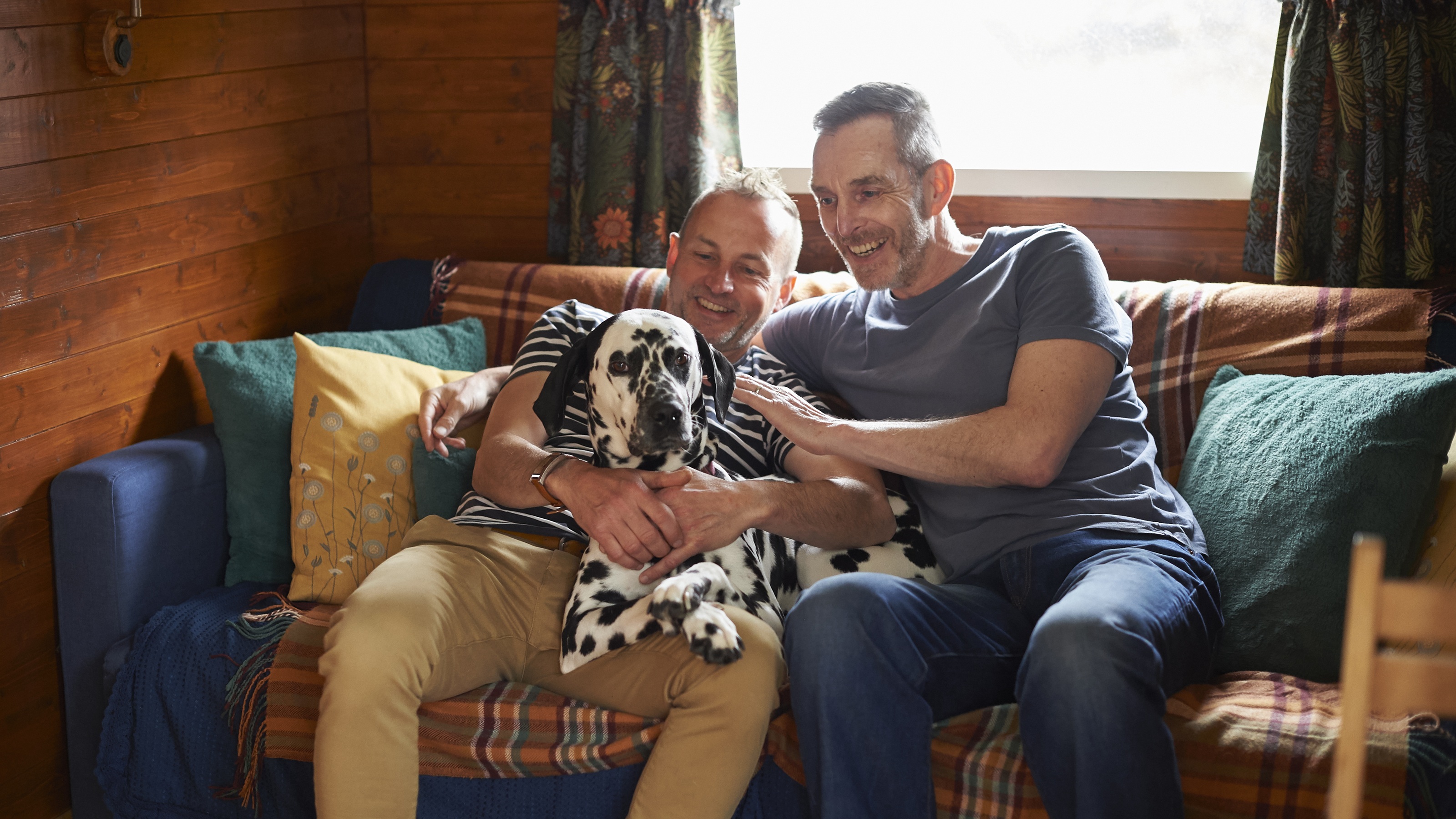 A same-sex couple cuddle with their dog on the sofa at a vacation cabin.