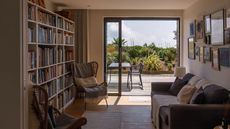 Sitting room with large, full bookshelf, two armchairs, gray sofa with cushions and throw, large and open sliding door revealing outdoor sitting area, greenery and blue sky with some clouds.