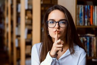 A women wearing glasses and smart attire holds her index finder against her lips in a "shh" gesture. Behind her, a library can be seen out of focus.