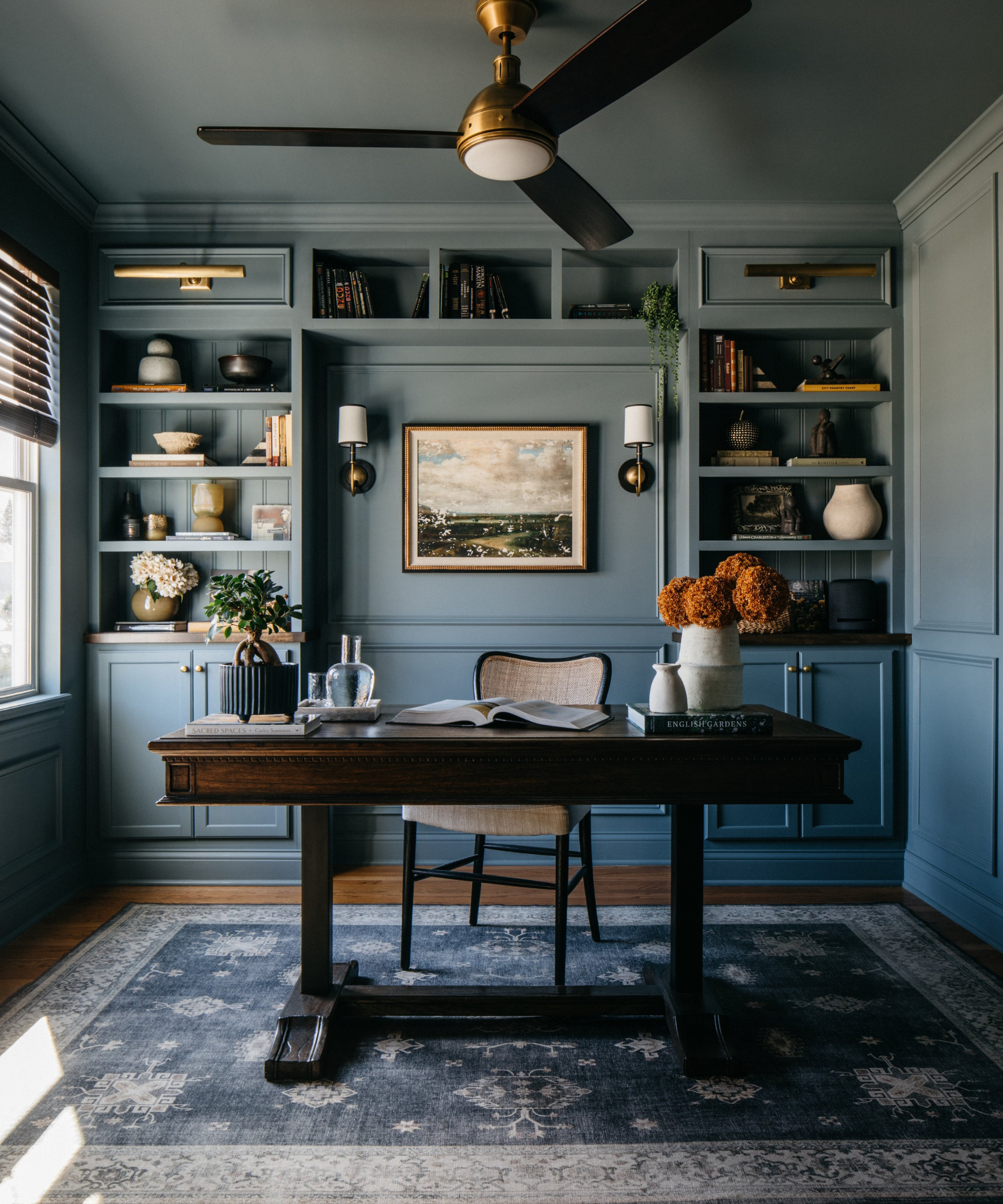 a home office drenched in a soft blue/gray color with built in shelves covered with decor, a freestanding desk with decor on top, a chair behind it, and a vintage-inspired rug on the floor