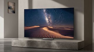 The Samsung R95H in a gray-colored living room displaying the Milky Way over sand dunes in the desert